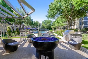 A fire pit sits in the middle of a patio with seating and a pool in the background